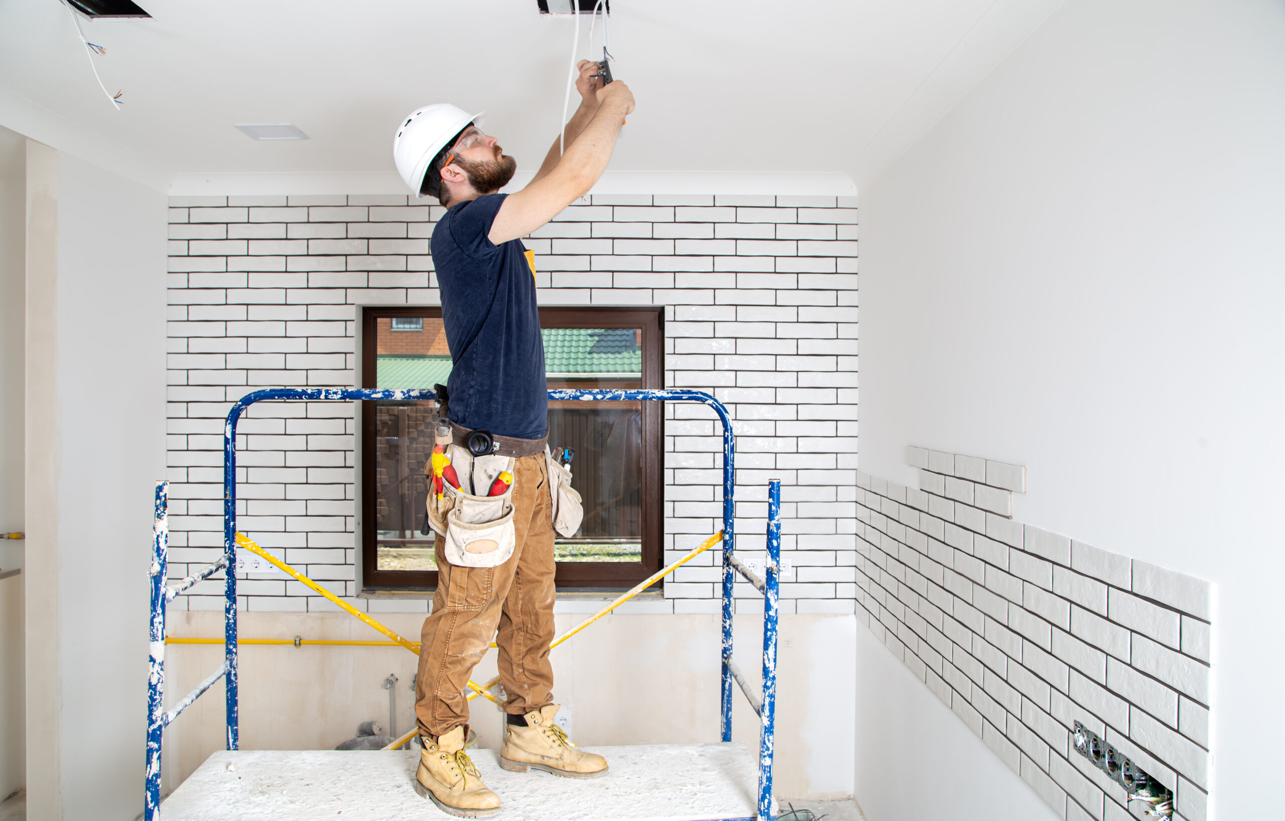Electrician Builder at work, installation of lamps at height. Professional in overalls with tools on the background of the repair site. Home renovation concept.